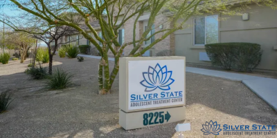 A sign reading Silver State Adolescent Treatment Center with a blue lotus logo and the address 8225 stands outside a beige building, surrounded by desert landscaping and trees—offering hope for teen mental health.