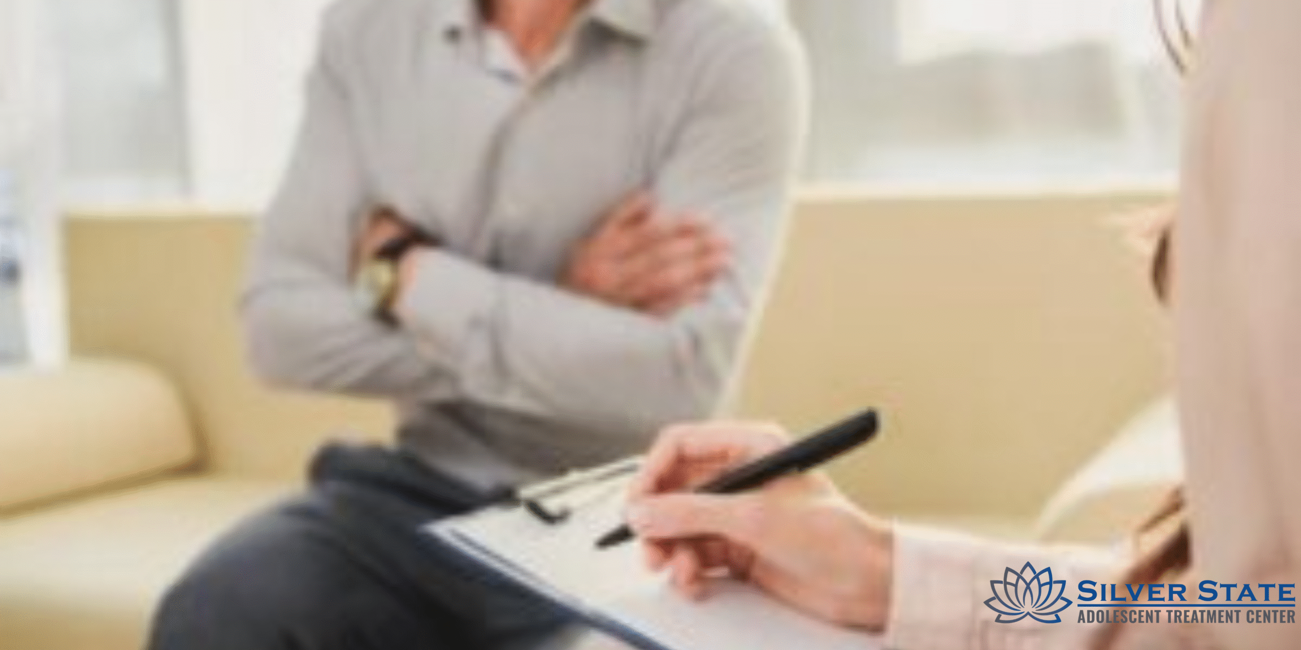 A person holding a clipboard and pen speaks with a teen who sits with crossed arms, highlighting the focus on adolescent mental health. The logo for Silver State Adolescent Treatment Center appears in the bottom right corner.