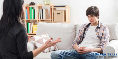 A teen boy sits on a couch looking thoughtful while listening to a woman, whose hands gesture as she speaks during a mental health counseling session. The room features shelves and home decor in the background.