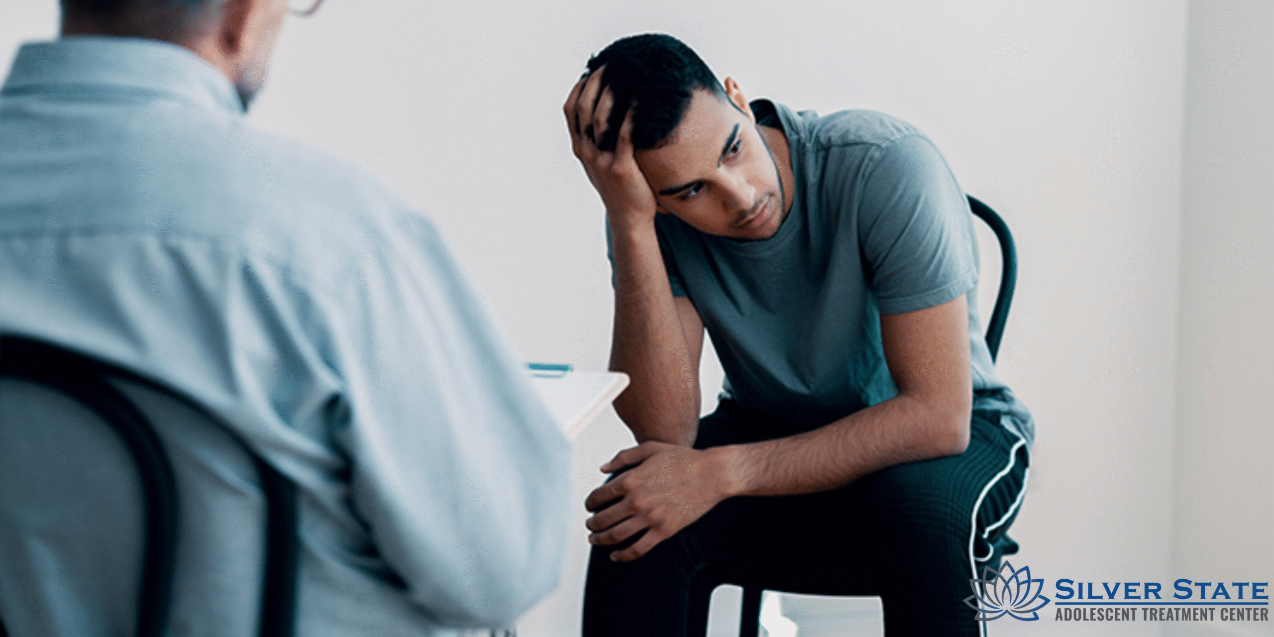 A distressed adolescent sits hunched forward, resting his head on his hand during a counseling session with a therapist. The Silver State Adolescent Treatment Center logo appears in the corner, highlighting a focus on teen mental health.