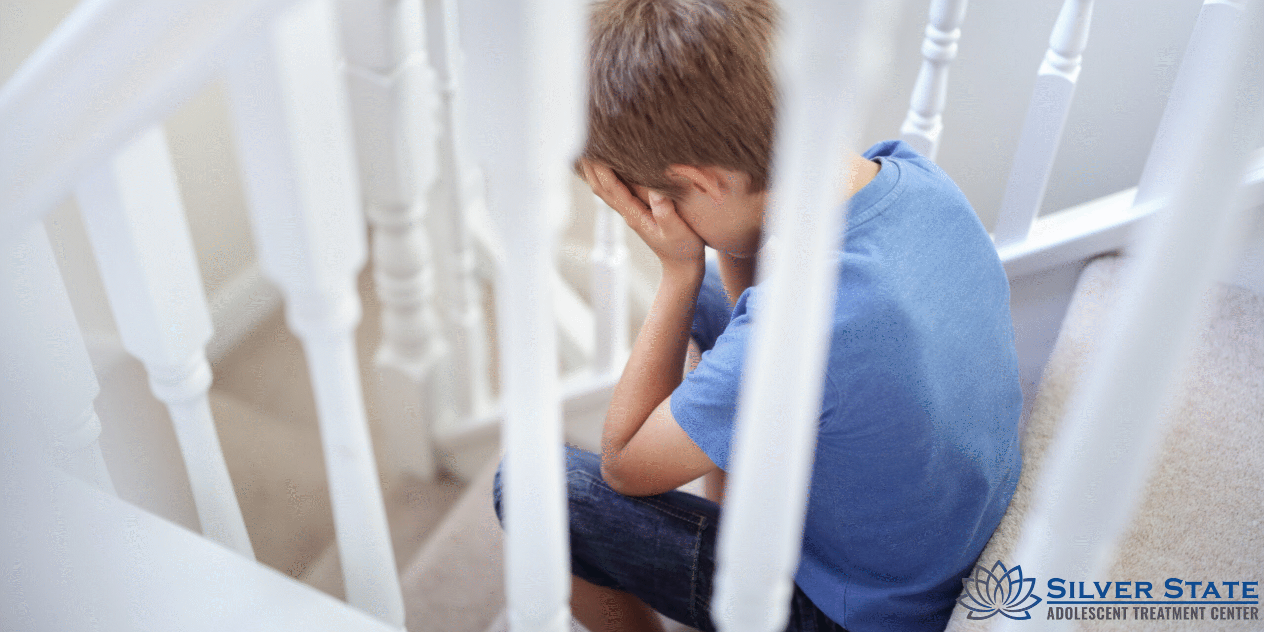 A teen in a blue shirt sits on a staircase with his face in his hands, appearing upset. The logo for Silver State Adolescent Treatment Center is visible in the bottom right corner, highlighting their focus on adolescent mental health.