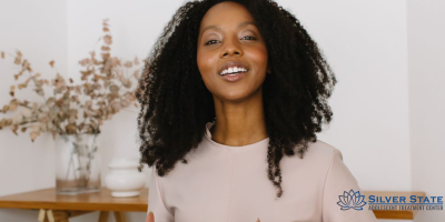 A woman with curly hair smiles while standing indoors near a wooden shelf with vases of dried flowers, reflecting a warm and supportive space for adolescent mental health. The Silver State Adolescent Treatment Center logo is visible in the bottom right corner.