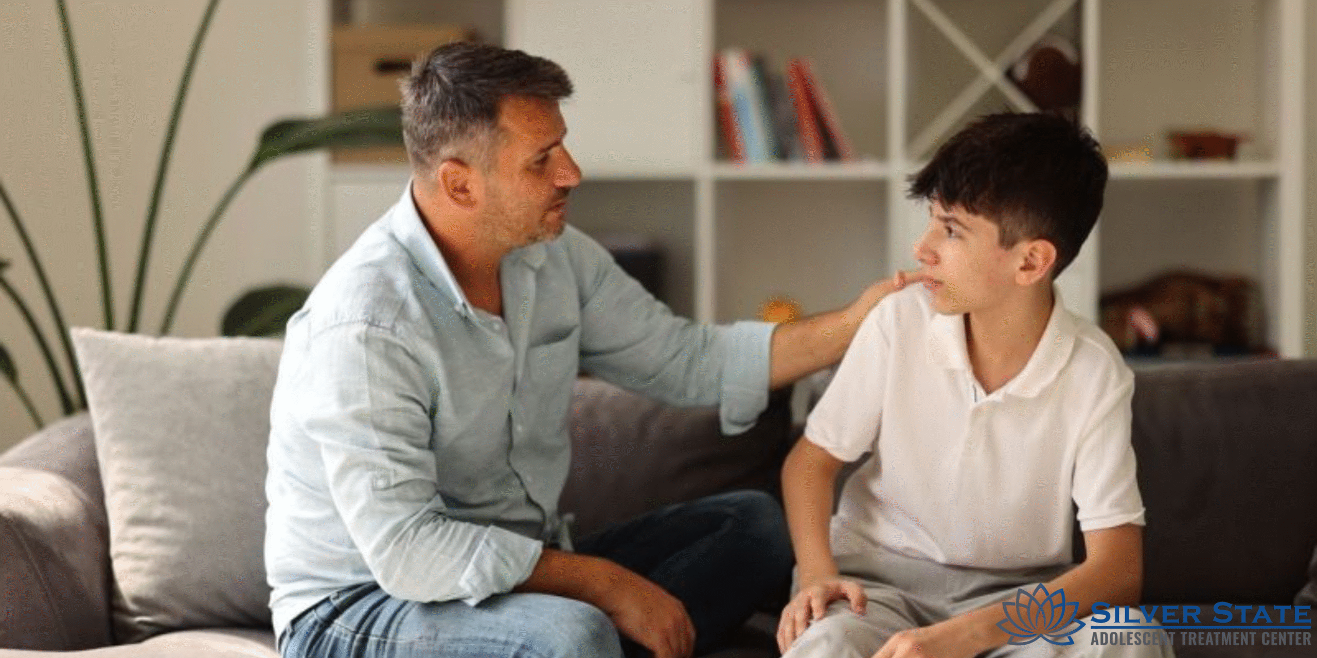 A man sits on a couch, gently placing his hand on the shoulder of a teen, offering comfort and support. Both appear to be engaged in a serious conversation about mental health in a home setting.