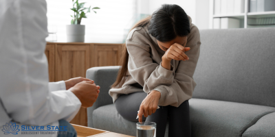 A young adolescent woman sits on a couch, leaning forward and wiping tears from her face with her arm. A person in a white coat sits nearby, suggesting a mental health therapy session. The Silver State Adolescent Treatment Center logo is visible.