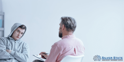 A teenage boy in a gray hoodie sits with crossed arms, looking at an older man with a notepad, suggesting an Alcohol Addiction Rehabilitation counseling session. The Silver State Adolescent Treatment Center logo is visible in the lower right corner.