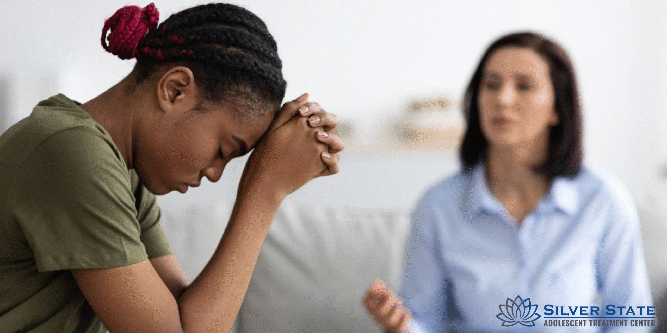A young adolescent woman sits with her head bowed and hands clasped, appearing distressed, while a therapist speaks to her during a mental health counseling session. The Silver State Adolescent Treatment Center logo is visible in the corner.