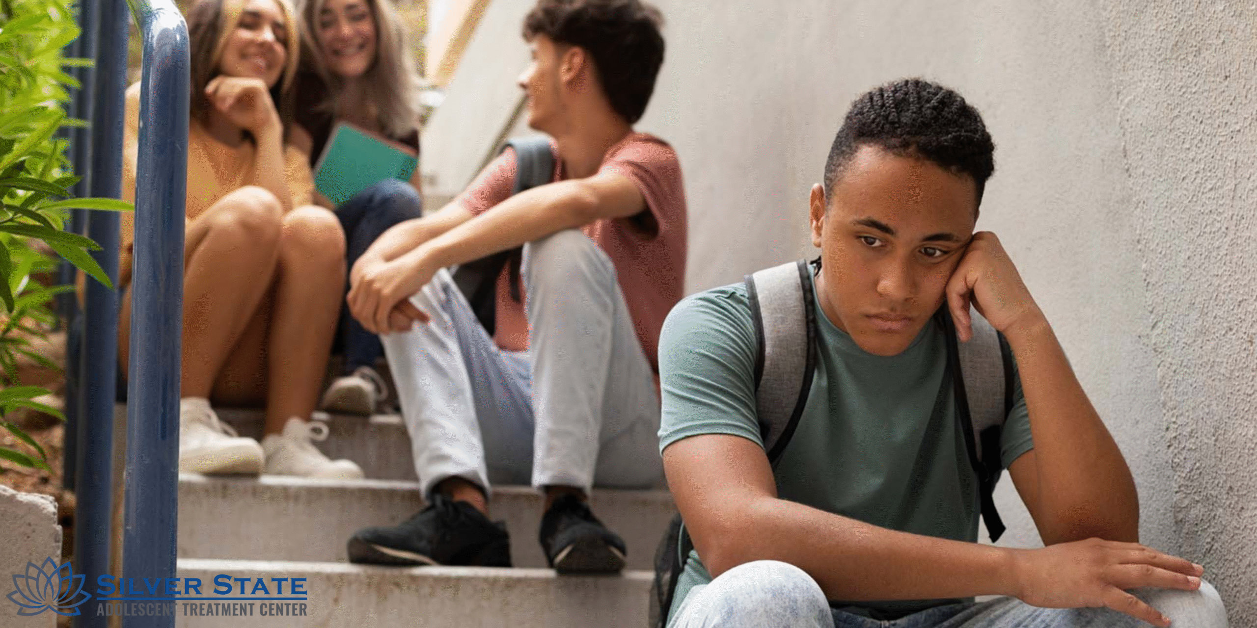 A sad adolescent boy sits alone on the stairs, his head resting on his hand, while three other teens chat and smile together in the background, highlighting the impact of social exclusion on teen mental health.